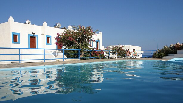 Une piscine à l’eau d’un bleu cristallin, un bâtiment blanc orné de motifs bleus, et quelques personnes assises au bord de la piscine.