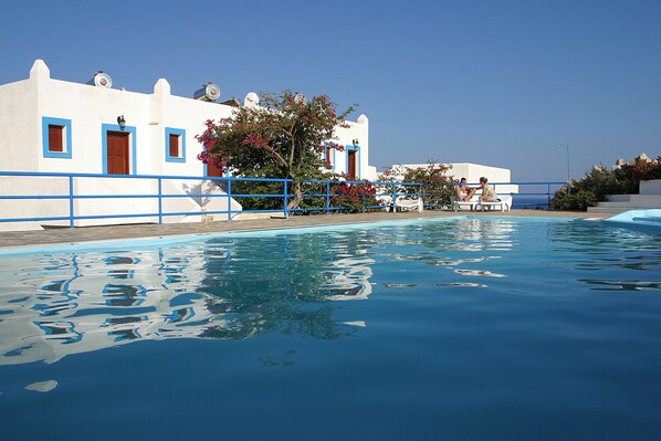 A swimming pool with clear blue water, a white building with blue trim, and a few people sitting by the pool.