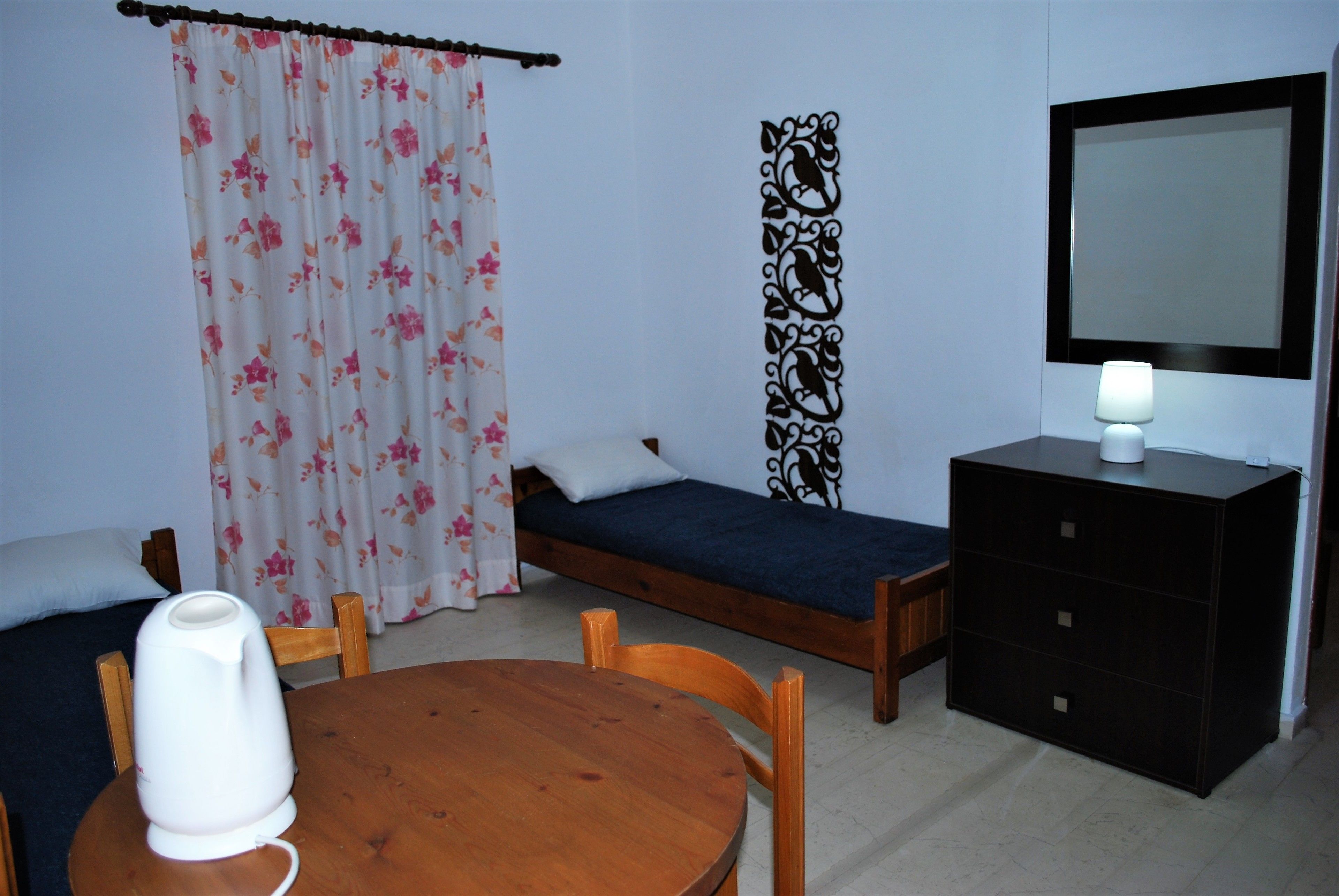 A room with a bed, a bedside table with a lamp, a wooden chair, a small table, and a curtain with a floral pattern.