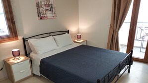 A bedroom with a bed, bedside tables, a lamp, a poster on the wall, and a view through a window. - Koula Apartments (Hersonissos)