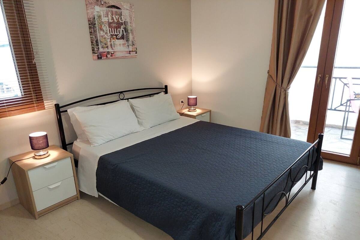 Ein Schlafzimmer mit Bett, Nachttischen, einer Lampe, einem Poster an der Wand und Blick durch ein Fenster.
