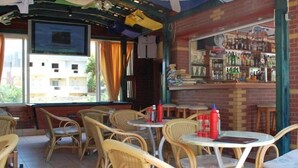 A bar area with wicker chairs and tables, a bar counter with bottles, and a view of buildings outside. - Koula Apartments (Hersonissos)