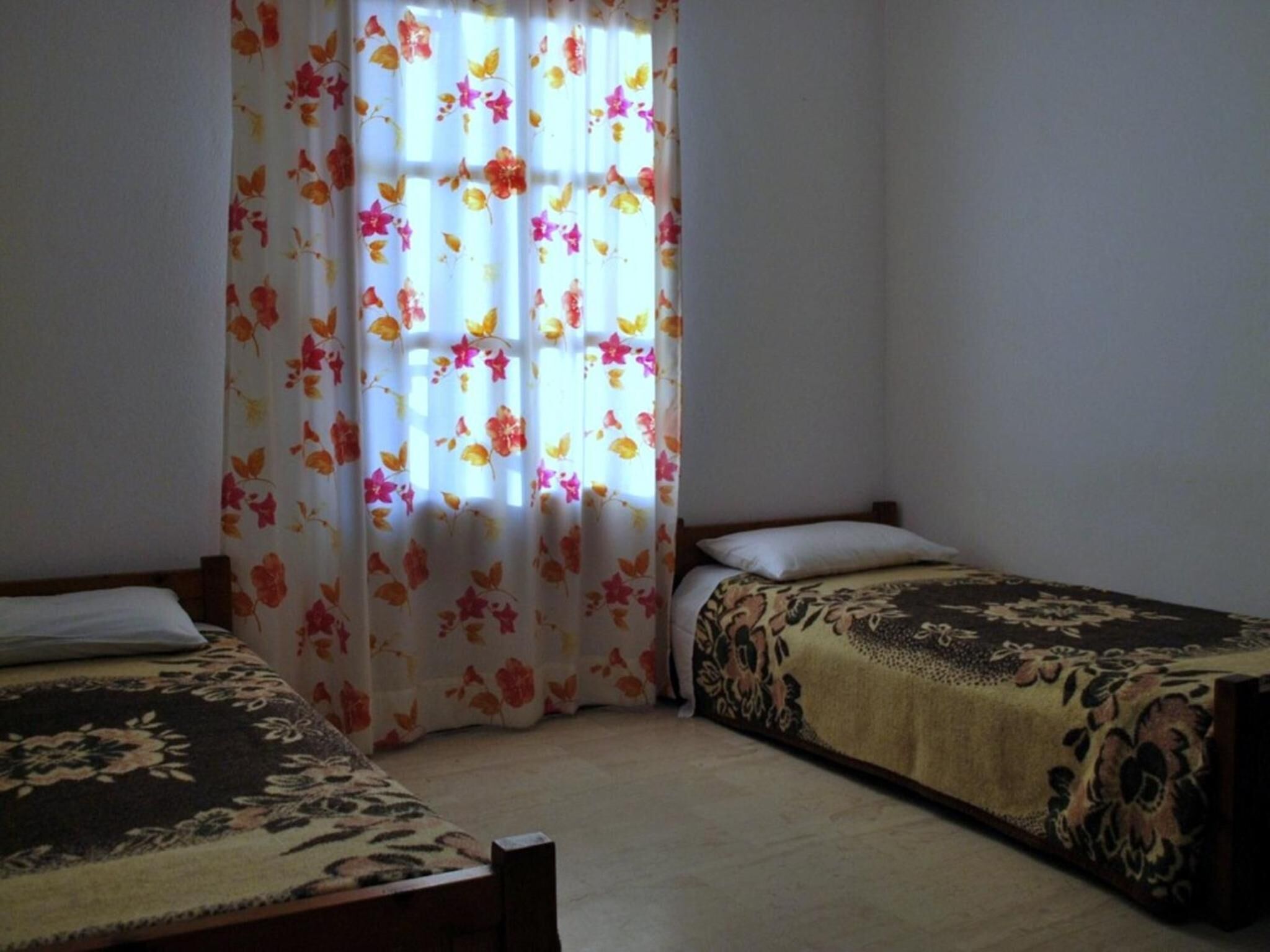A room with two single beds, a window with floral curtains, and a plain wall.