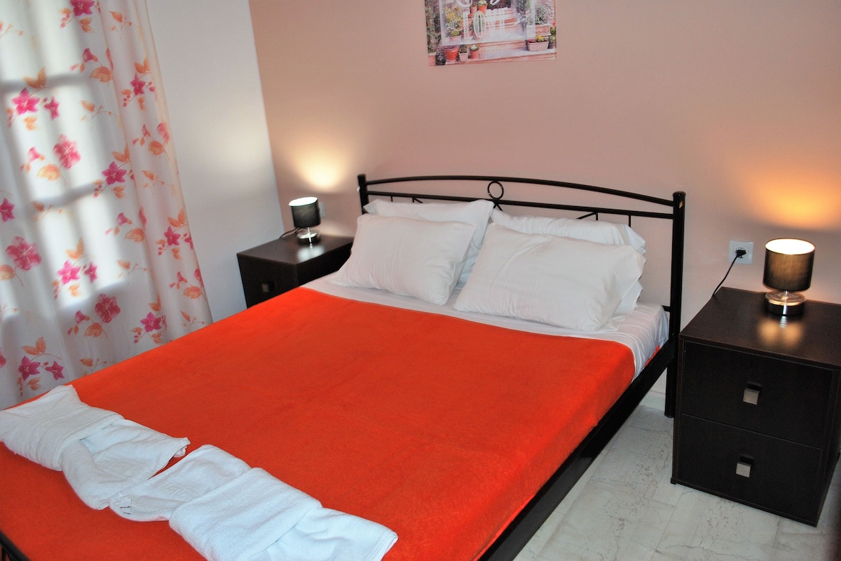 A neatly made bed with an orange bedspread, white pillows, and a folded white towel. There are two bedside tables with lamps, and a wall-mounted picture above the bed.
