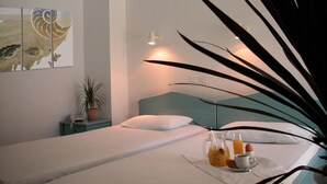 Una cama bien tendida con un almohadón blanco y un cabecero color turquesa, una mesita de noche con una planta en maceta y una bandeja de desayuno con jugo de naranja, huevos y una taza.