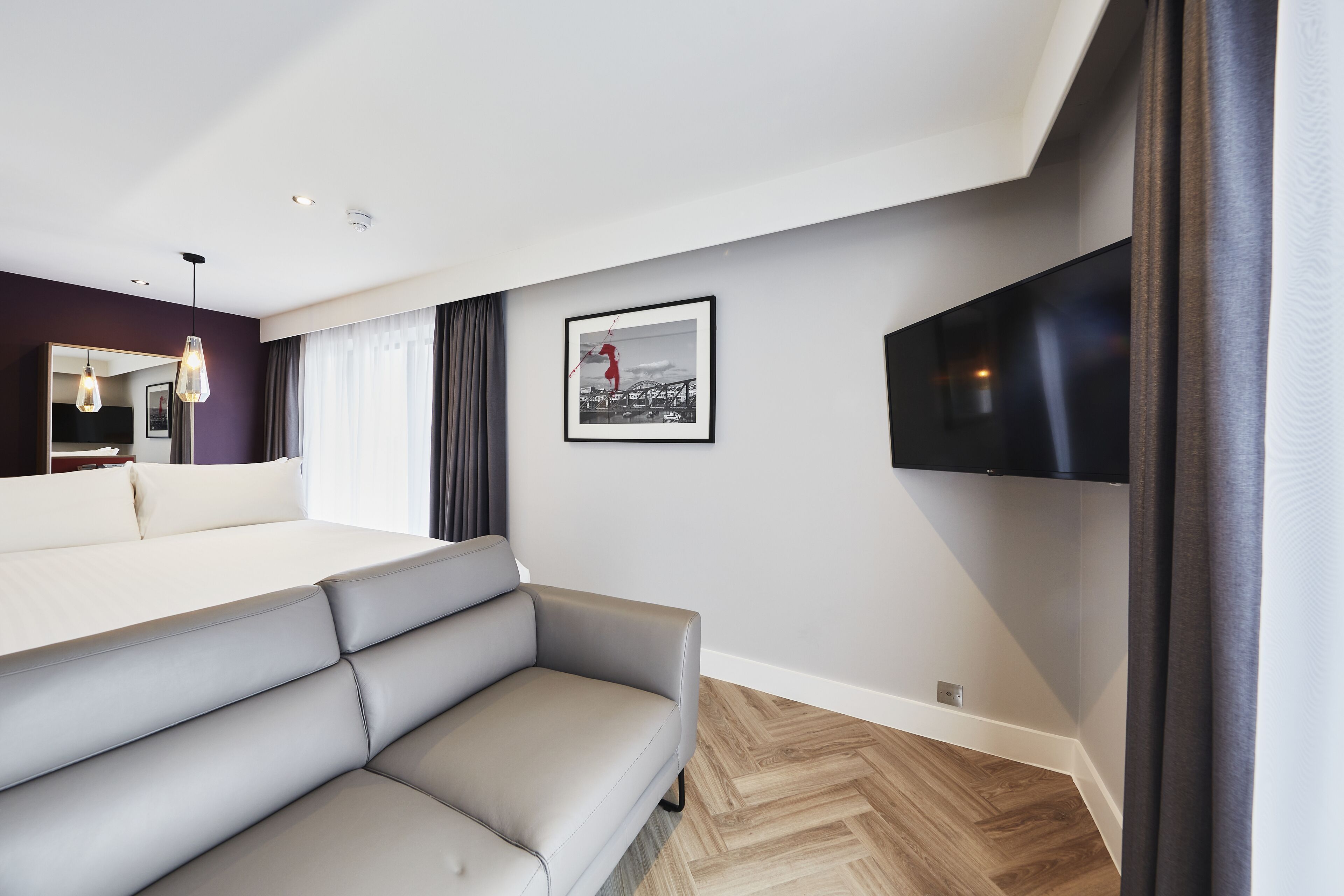 A modern hotel room with a sofa, a flat-screen TV, a framed picture on the wall, and a large window with curtains.