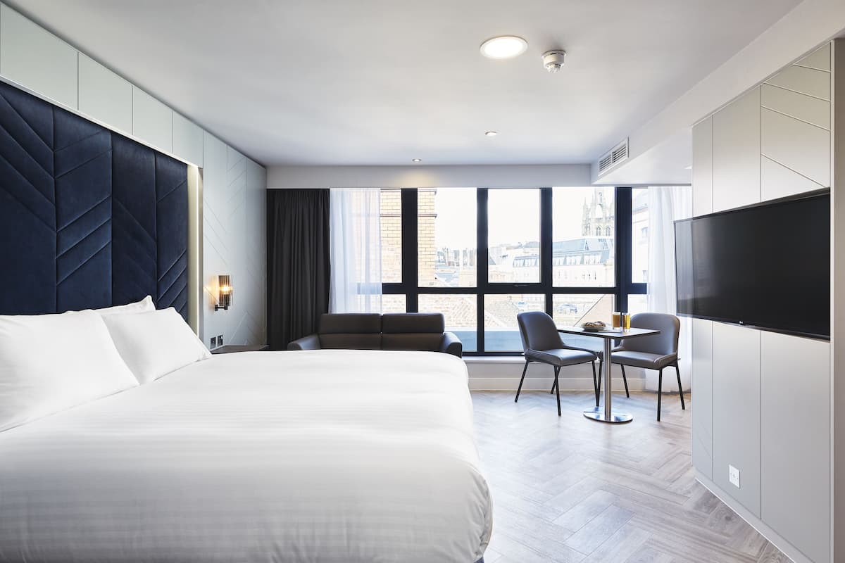 A modern hotel room with a large bed, a desk with two chairs, a flat-screen TV, and a city view.