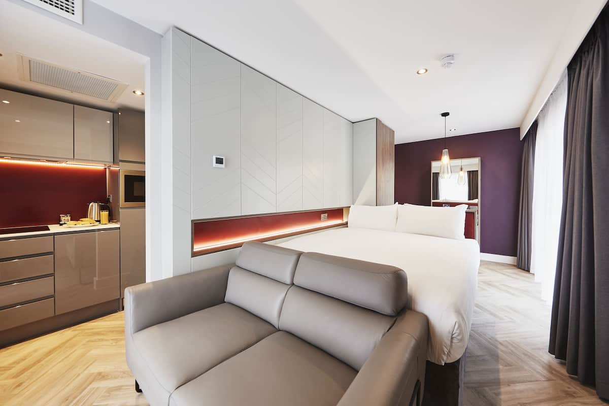 A modern hotel room with a sofa, a bed, a kitchenette, and a large mirror.