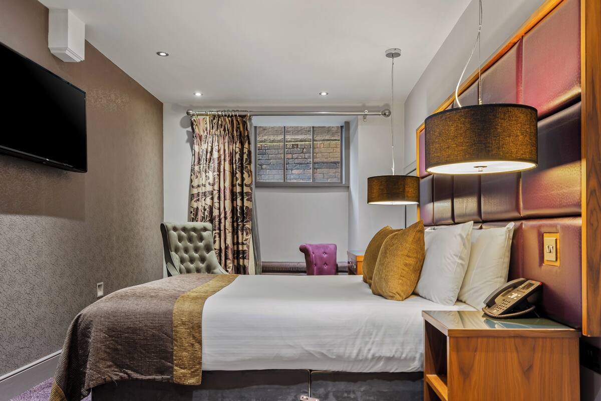 A hotel room with a bed, a television, a window with curtains, a lamp, and a telephone on the nightstand.