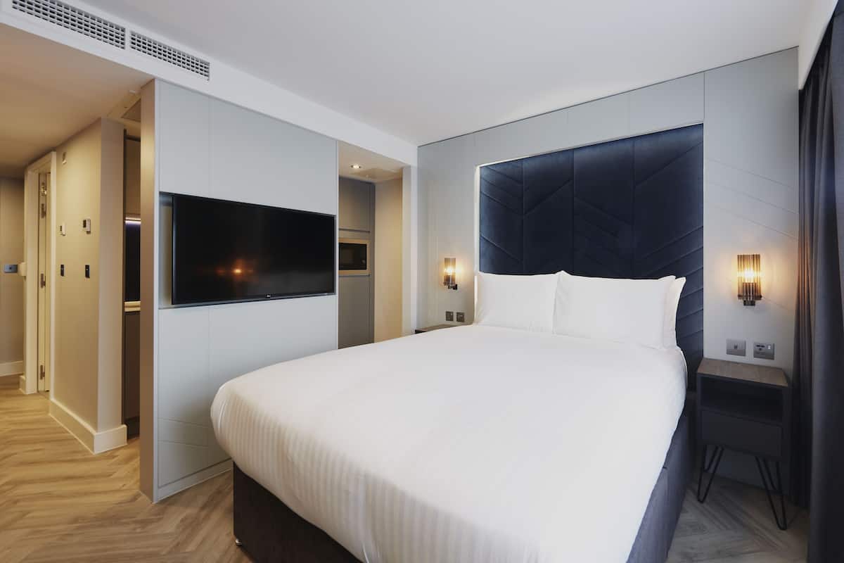 A modern hotel room with a large bed, a flat-screen TV mounted on the wall, and a small bedside table with a lamp.