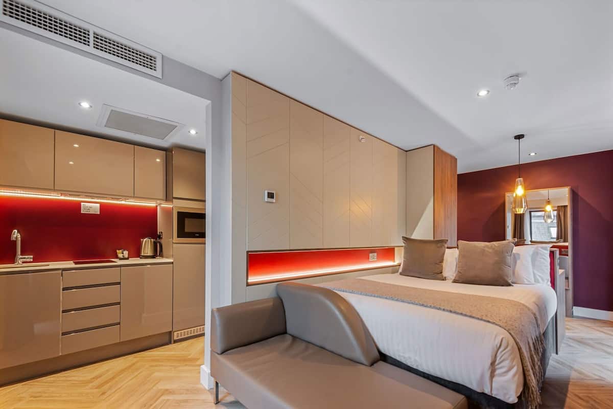 A modern hotel room with a kitchenette, a bed with a headboard, and a seating area.