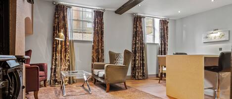 A modern living room with a sofa, armchairs, a glass coffee table, and a wooden floor. There is a desk with a chair and a bar stool. The room has patterned curtains and a ceiling with exposed beams.