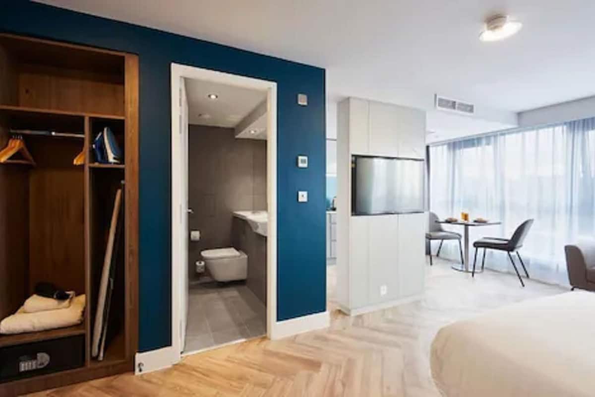A hotel room with a wooden wardrobe, a bathroom with a toilet and sink, a dining area with a table and chairs, and a TV mounted on the wall.