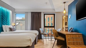 A hotel room with two beds, a desk, and a chair. There is a large window with a view of buildings outside. - Roomzzz Newcastle (Newcastle-upon-Tyne)