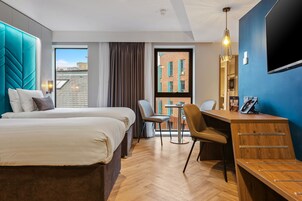 A hotel room with two beds, a desk, and a chair. There is a large window with a view of buildings outside.