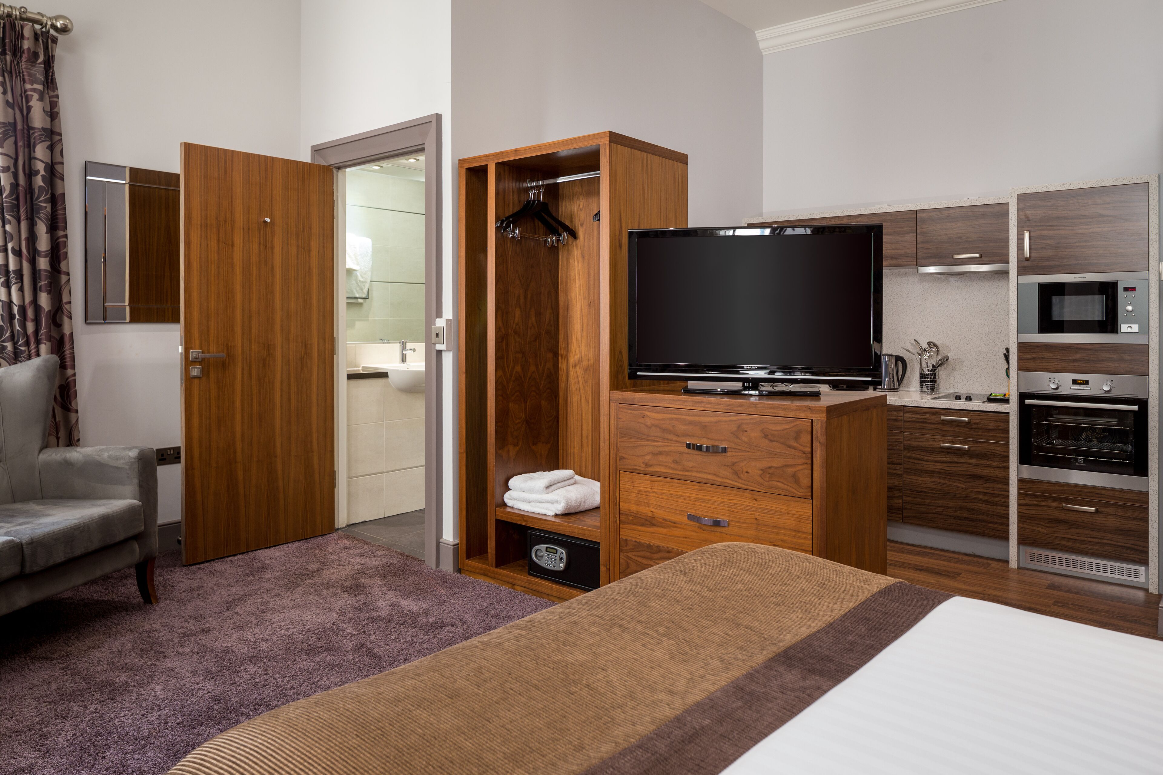 A hotel room with a bed, a TV, a wooden wardrobe, a grey armchair, and a kitchenette with a microwave and oven.