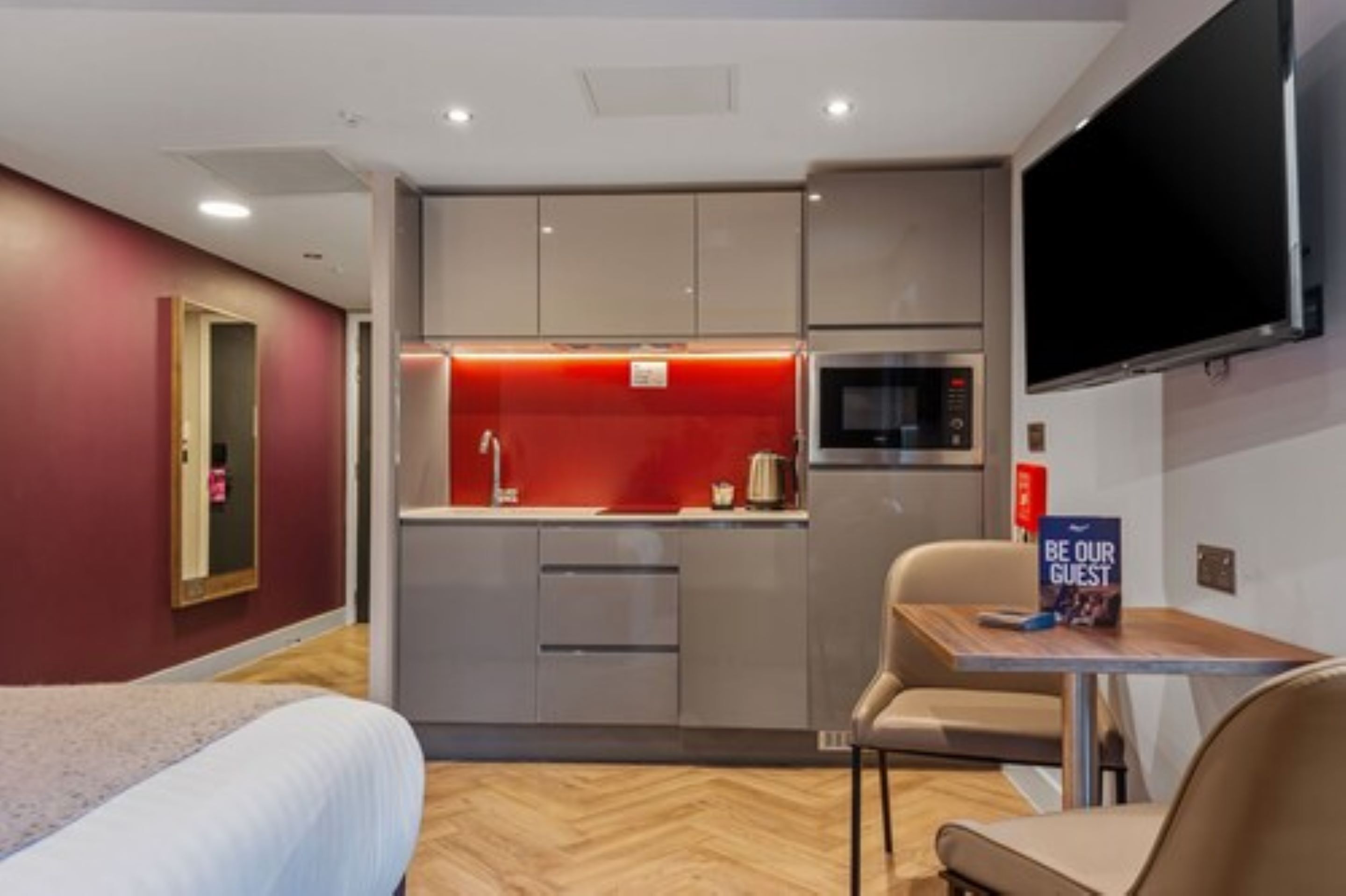A compact hotel room with a kitchenette, a bed, a TV, and a small table with a 'Be Our Guest' sign.