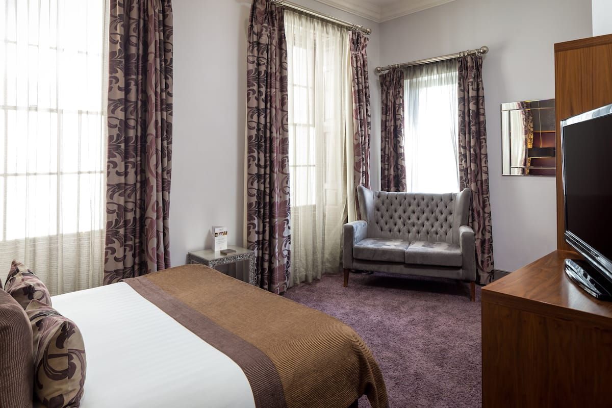 A hotel room with a large bed, a grey armchair, a wooden TV stand, and a window with patterned curtains.