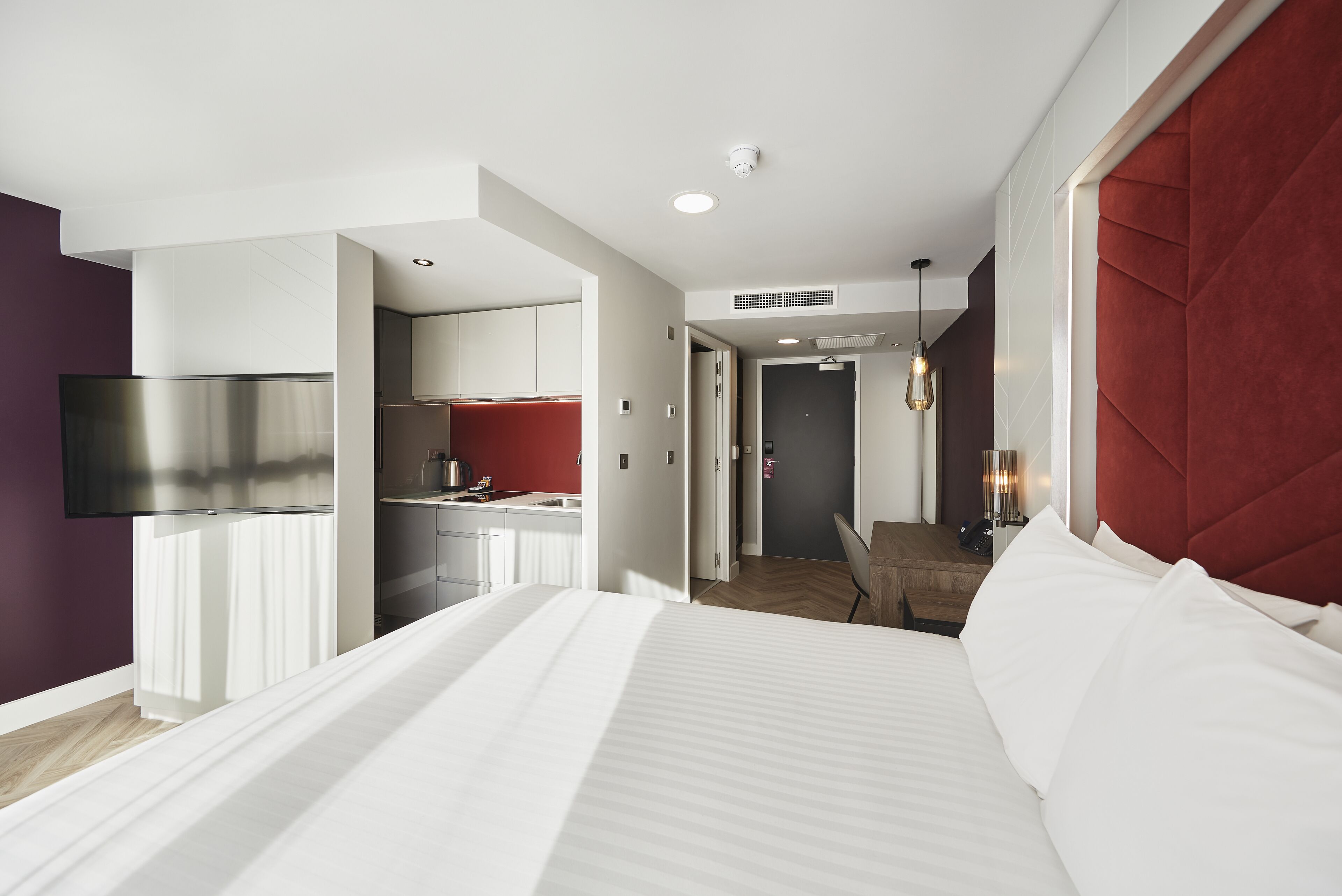 Une chambre d’hôtel moderne dotée d’un grand lit, d’un téléviseur fixé au mur, d’une kitchenette et d’un mur d’accent de couleur rouge.
