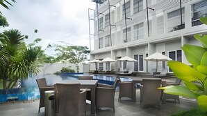 Un espace extérieur avec piscine, des chaises longues et des parasols, entouré de verdure et bordé d’un bâtiment moderne doté de balcons.