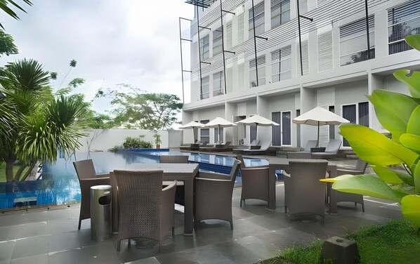 An outdoor pool area with lounge chairs and umbrellas, surrounded by greenery and a modern building with balconies. - Grand Kecubung Hotel (Pangkalan Bun)