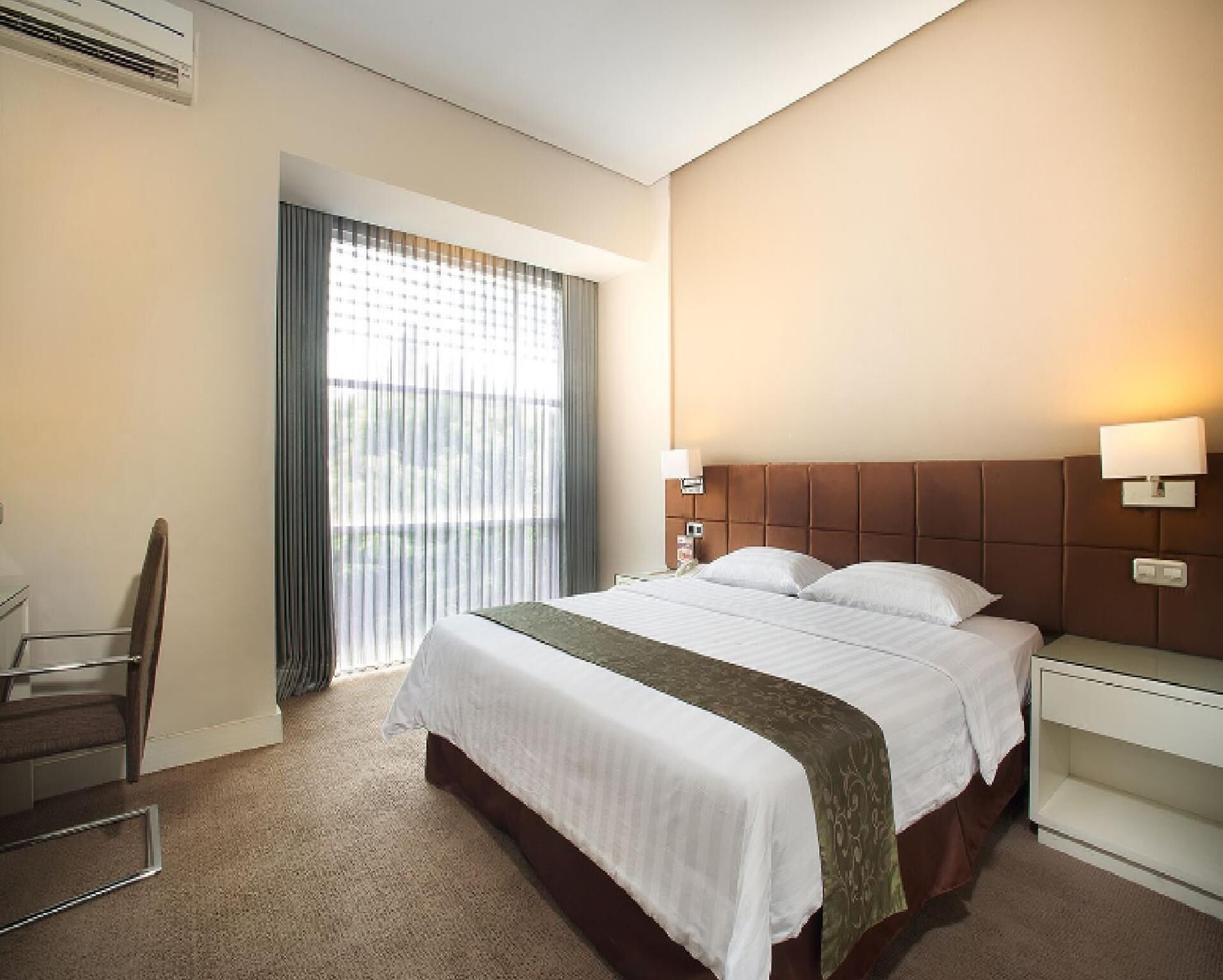 A hotel room with a bed, a desk, a chair, a window with curtains, and a wall-mounted lamp.