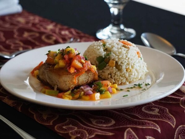 A plate with a chicken dish and rice, accompanied by a glass and spoon on a table with a patterned tablecloth. - Grand Kecubung Hotel (Pangkalan Bun)