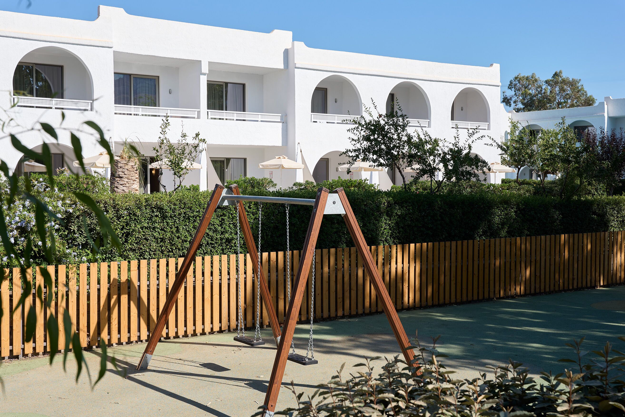 A white building with multiple balconies and arched windows, a wooden fence, and a swing set.