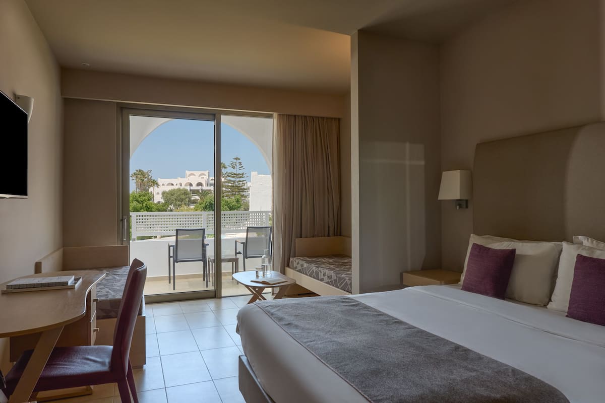 A hotel room with a bed, a desk, a chair, a small table, and a balcony with a view of buildings.