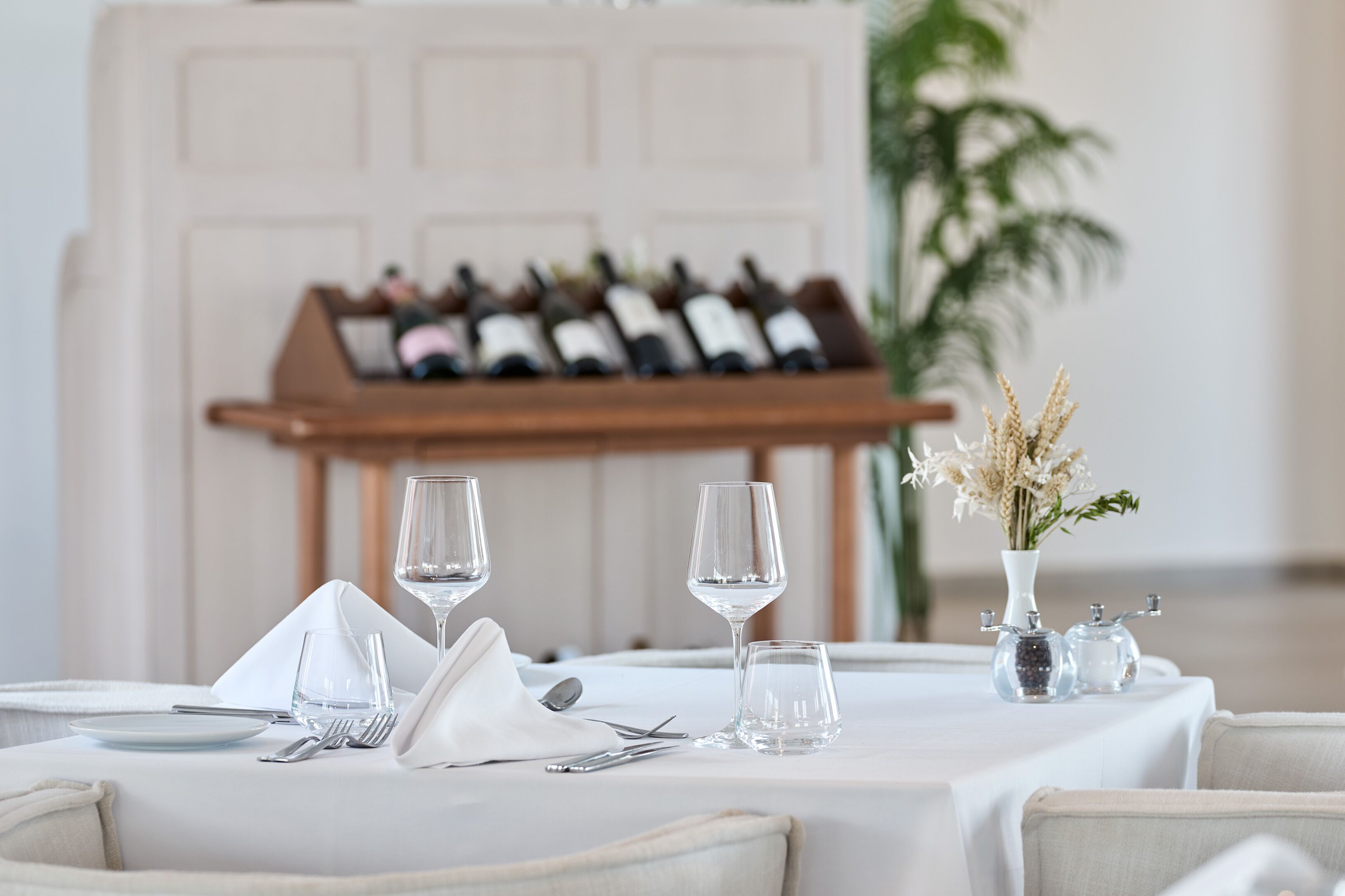 Une table à manger dressée avec des verres à vin, un vase contenant des fleurs séchées et un meuble à bouteilles de vin en arrière-plan.