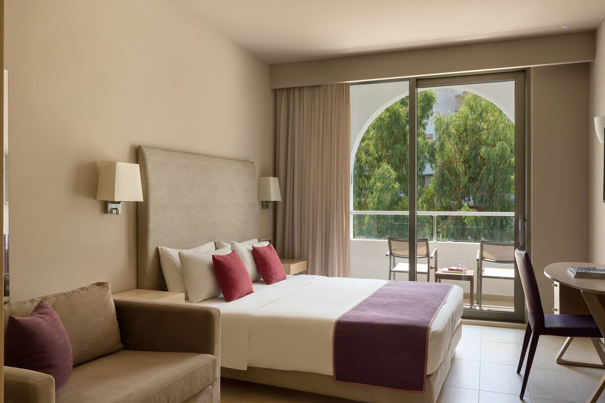 A hotel room with a bed, a sofa, a balcony with a table and chairs, and a view of trees.
