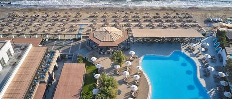 An aerial view of a beach resort with a swimming pool, beach umbrellas, lounge chairs, and a covered seating area.
