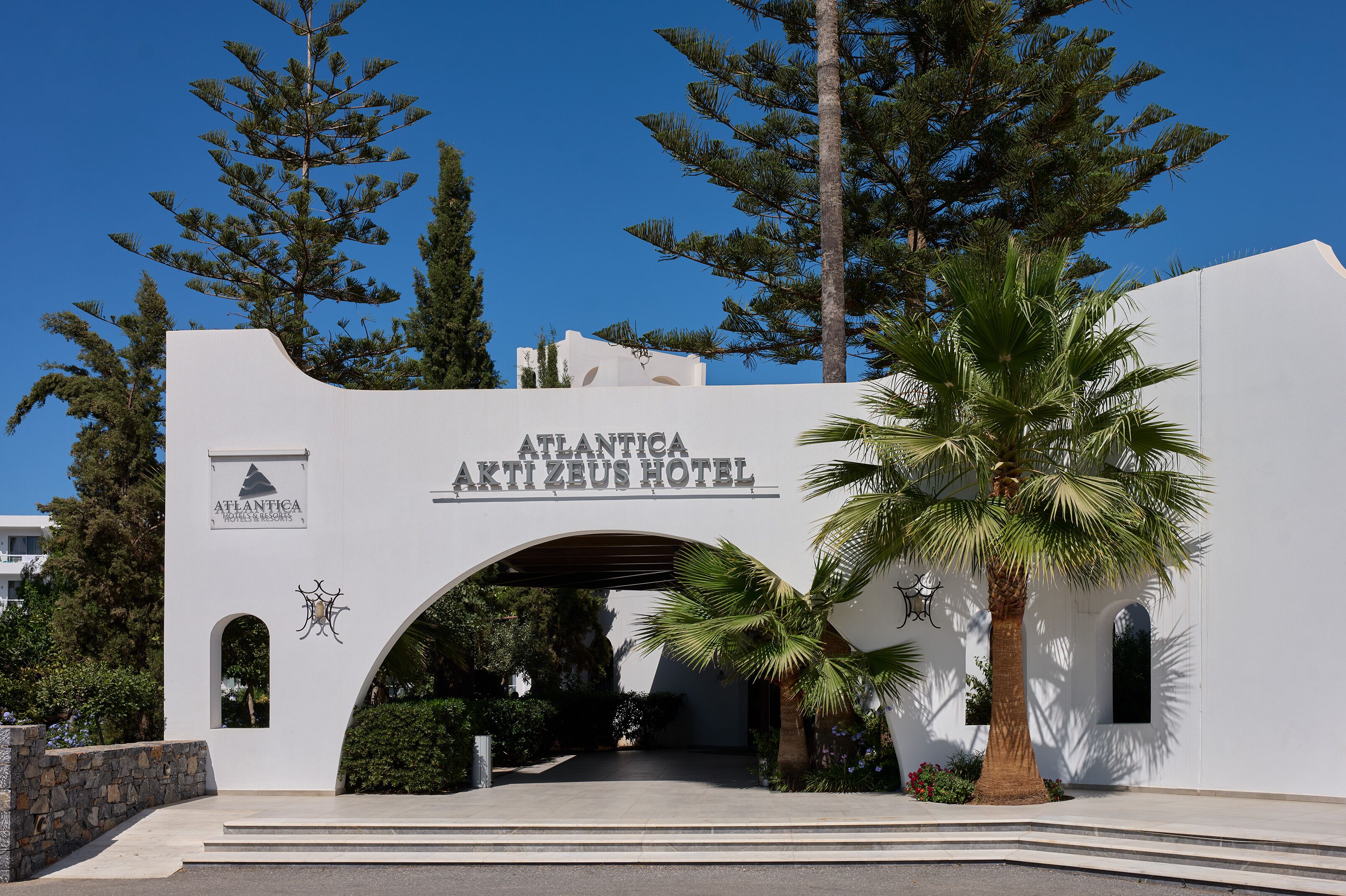 The entrance to the Atlanticia Akti Zeus Hotel with a clear archway and a palm tree in the foreground.