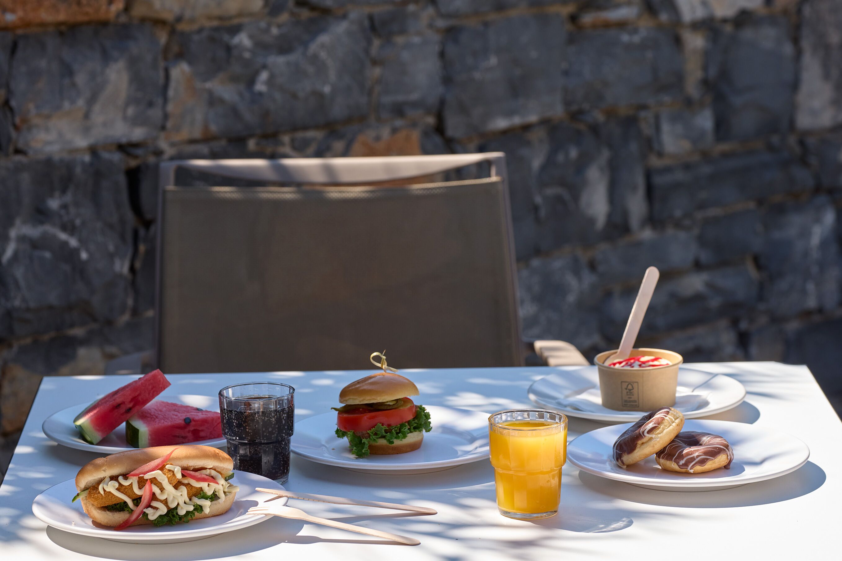 A table with a sandwich, watermelon slices, a glass of juice, and a bowl of yogurt with a spoon.