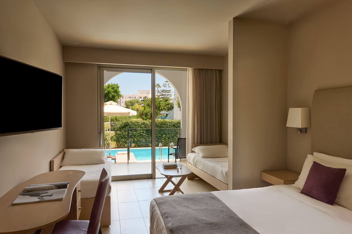 A hotel room with a bed, a desk, a chair, a small table, a TV, and a view of a pool and greenery.