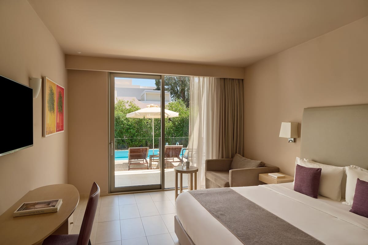 A hotel room with a bed, a desk, a chair, a TV, and a sliding glass door leading to an outdoor pool area.