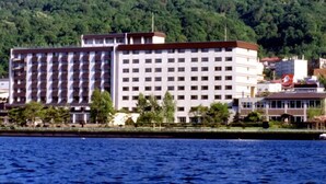 Un hotel de varios pisos con fachada simétrica, situado cerca de una masa de agua y un paisaje montañoso cubierto de bosque al fondo.