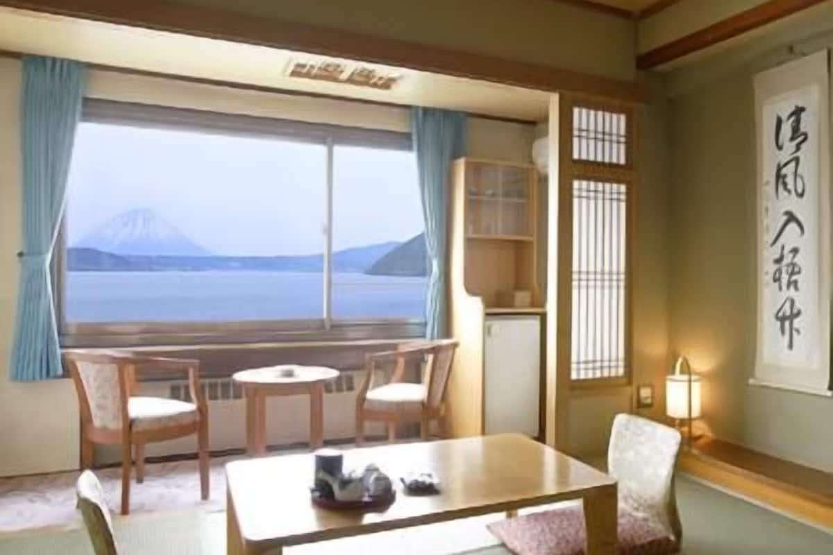 Una habitación japonesa tradicional con suelo de tatami, una mesa baja, sillas, una mesita, una lámpara y una vista de la montaña a través de una ventana.