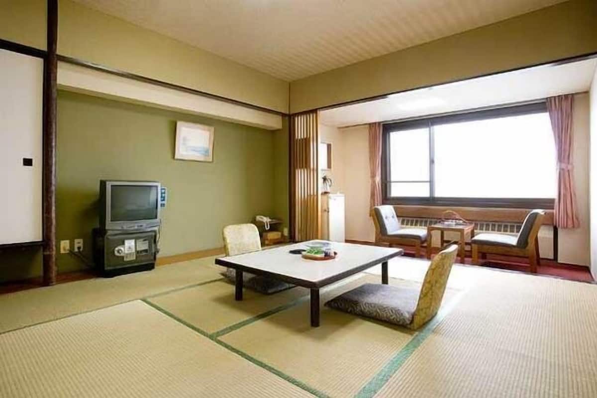 Una habitación japonesa tradicional con suelo de tatami, un televisor, una mesa y vistas al exterior.