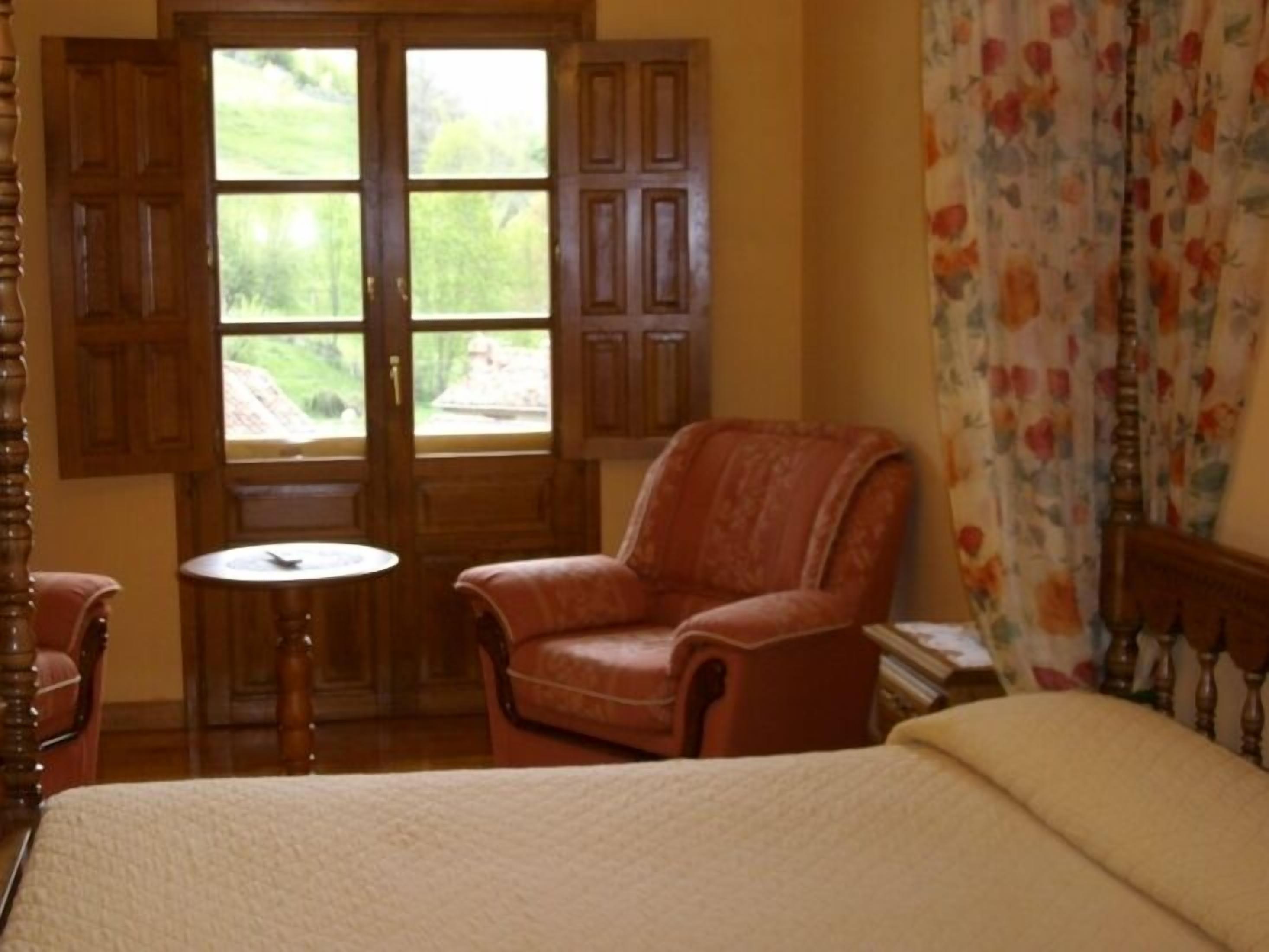 A room with a bed, a wooden chair, a small round table, and a window with wooden shutters.