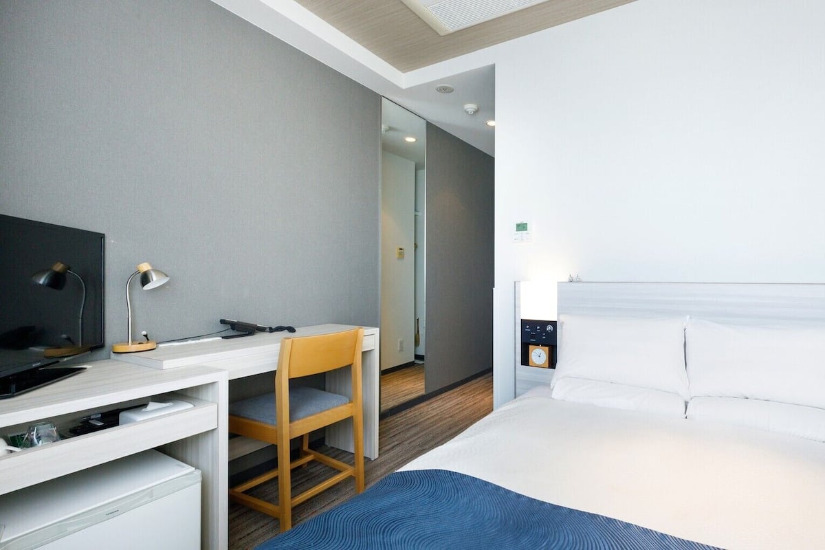 A hotel room with a bed, a desk with a chair, a television, and a clock on the wall.