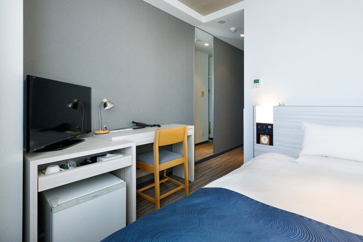A hotel room with a bed, a desk with a TV, a chair, a small desk, a clock, and a window.