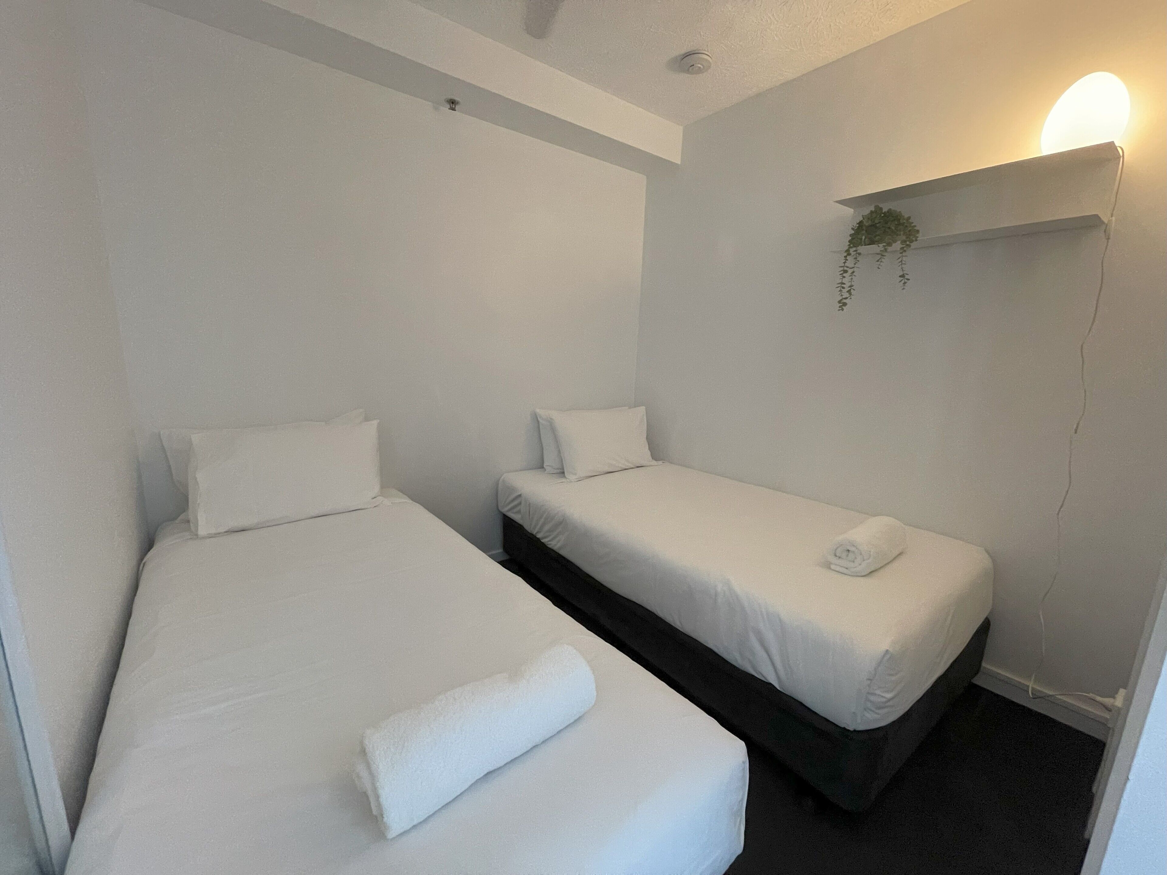 Quarto pequeno e limpo com duas camas individuais, uma estante na parede com uma planta e uma luminária no teto.