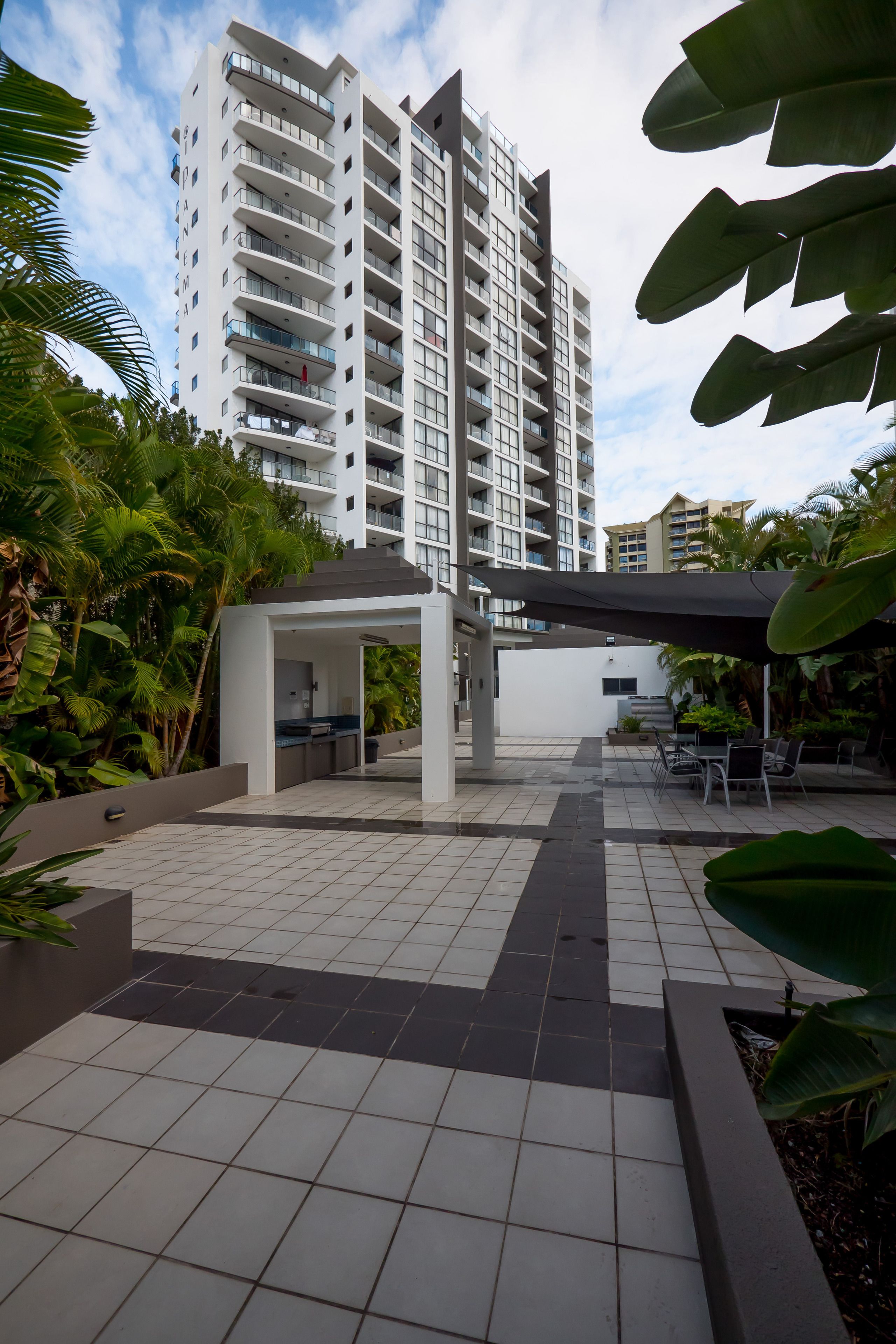 A modern outdoor patio area with a covered seating area, surrounded by lush greenery and a tall residential building in the background.