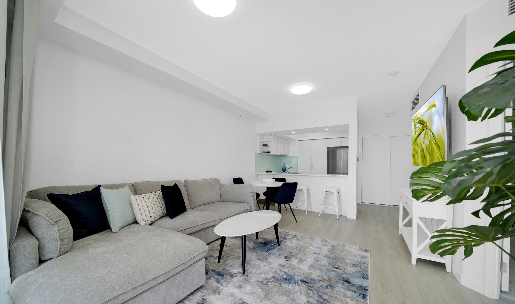 A modern living room with a grey sofa, a white coffee table, and a blue and white rug. There is a kitchen area in the background with white cabinets and a stainless steel refrigerator.