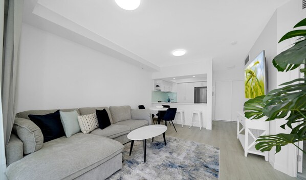 Una sala de estar moderna con un sofá gris, una mesa de centro blanca y un tapete azul y blanco. Se observa una cocina al fondo con armarios blancos y un refrigerador de acero inoxidable.