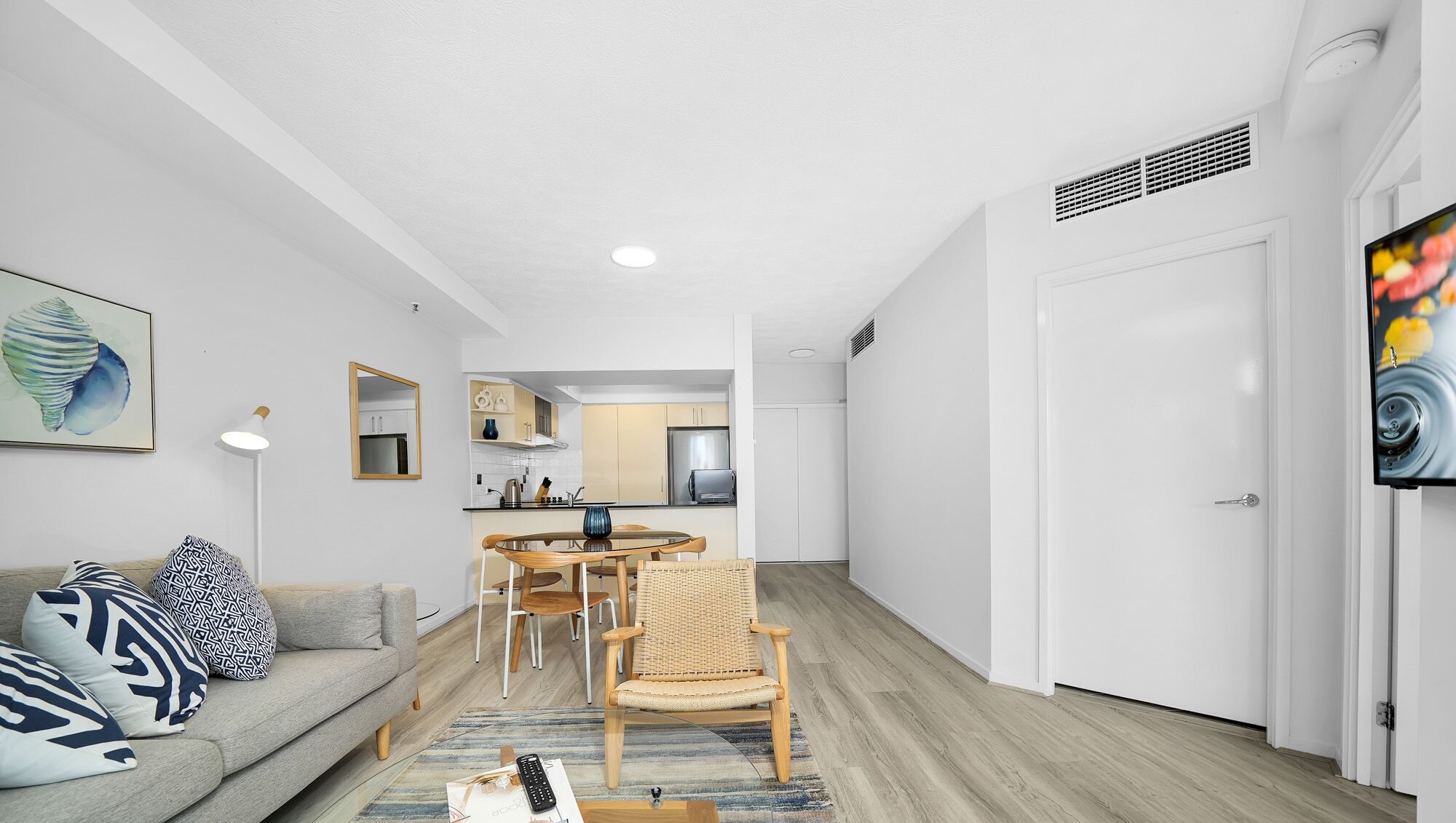 Una sala de estar moderna con un sofá, una mesa de comedor con sillas, una cocina al fondo y un televisor grande en la pared.