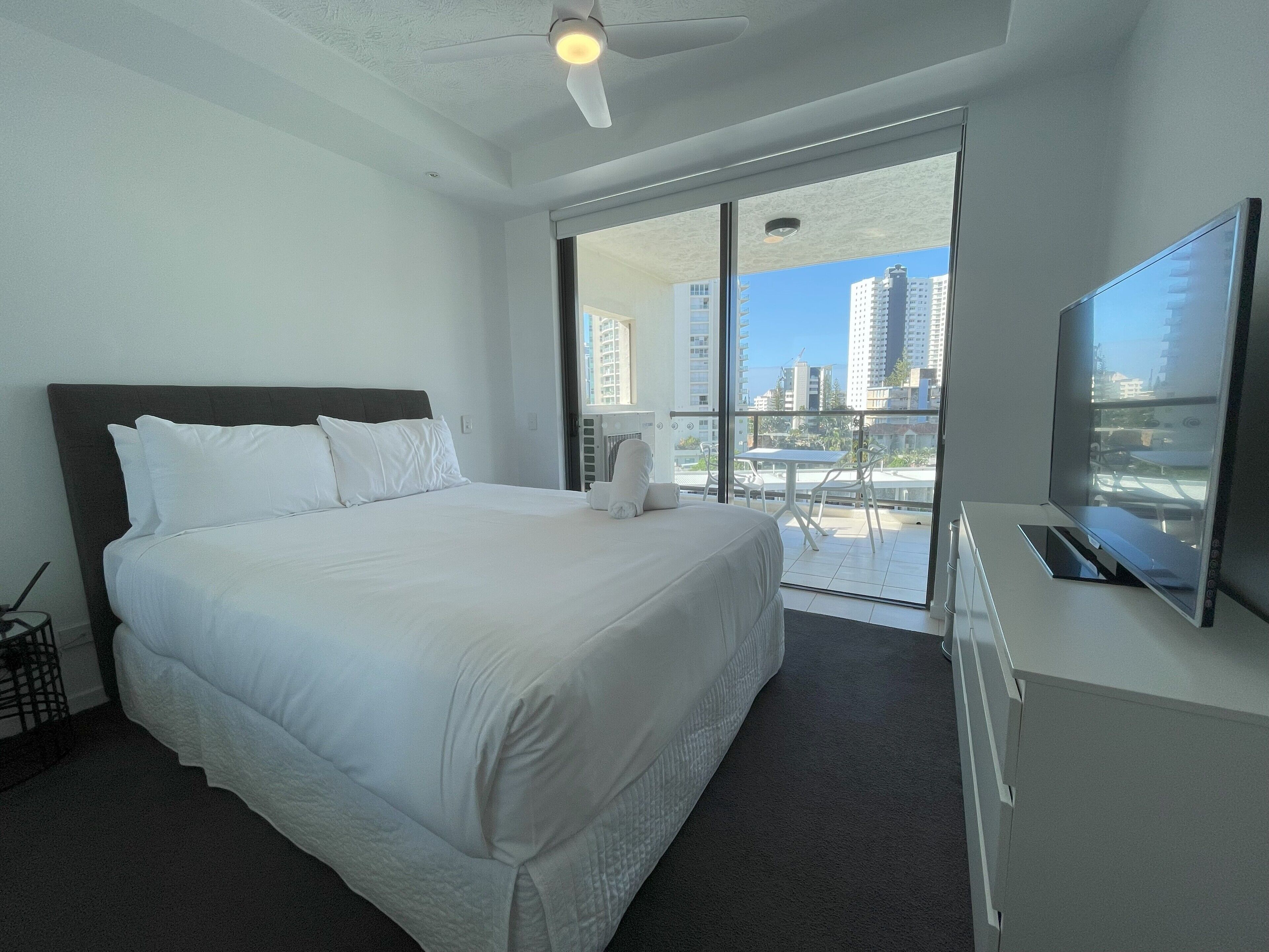 A hotel room with a large bed, a flat-screen TV, a ceiling fan, and a balcony with a view of tall buildings.