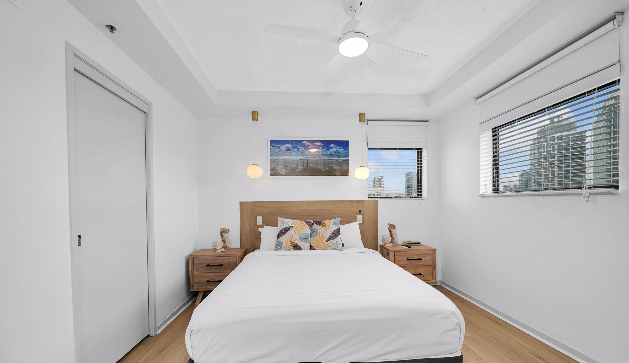 A bedroom with a bed, two bedside tables, a ceiling fan, a window with blinds, and a painting on the wall.