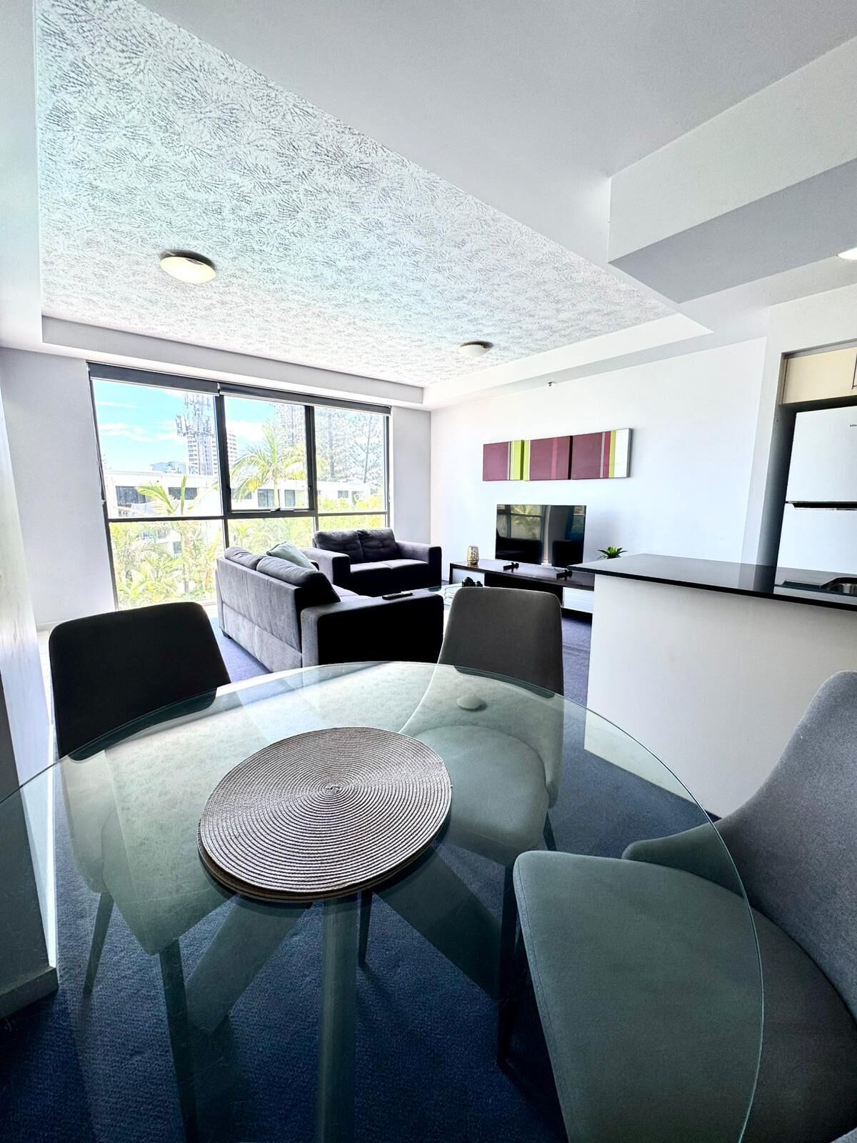 Un salon moderne avec une table en verre, des canapés gris et une grande fenêtre donnant sur la ville.